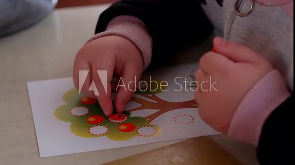 Child's hands applying red plasticine dots to a colorful tree illustration on a table, showcasing a creative activity for children using plasticine materials