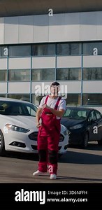 Automotive mechanic in red overalls stands outside a modern auto service center, surrounded by vehicles, demonstrating professionalism and customer engagement