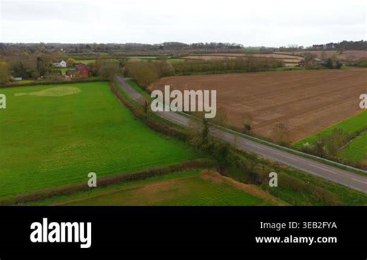 Rural road with several cars moving along farmland bordered by green fields and plowed soil under overcast sky. Countryside landscape with village houses and tree lines creating layered agricultural view. Transport and farming concept spring season Stock Video Footage - Alamy