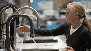 A woman with blonde hair in a grey shirt is examining a kitchen sink faucet display in a store. Shes touching the faucet with her hand, looking at it closely.