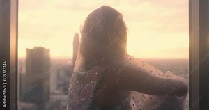 Beautiful young woman draped in translucent material sits silhouetted in an apartment window high above Downtown Los Angeles with sunset sky. Medium close up, recorded with black Promist filter.