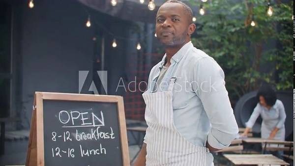 African American man writing on board inviting message. Friendly male owner welcoming new customers to try food of his cafe. In blurred background visible hardworking waitress cleaning terrace zone.