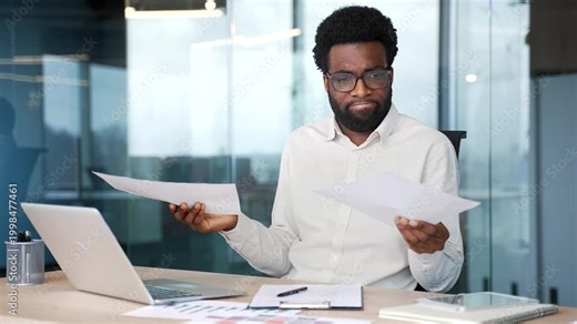 Confused puzzled african american businessman having difficulty with paperwork sitting at workplace in business office. Frustrated employee looks at the documents and cannot understand the problem