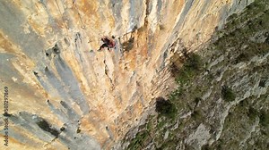 AERIAL: Sporty young woman top rope climbing up the picturesque limestone wall. Female rock climber on her way up the wall unhooking the rope. Adrenaline sport activities in breathtaking environment.