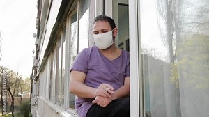 A man in a medical mask sits on a window frame, smokes a cigarette through a mask, puffs it twice and puts it out with his hands. medium shot, Full HD, no sound.