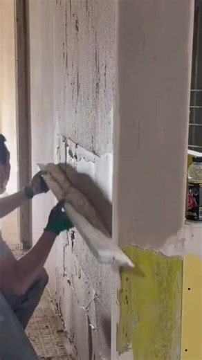 scraping old white peeling paint from an indoor vertical wall surface using a sharp metal scraper