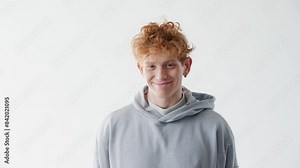 Young handsome Caucasian man with red hair and grey hoodie standing in front of white backdrop. Smiling while looking directly at camera. Male modelling. Happy expression during casting.
