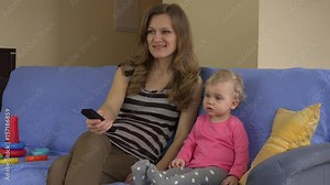 Mom with remote and daughter watching television sitting on sofa.