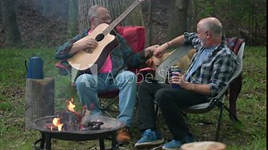Two gay men in front of campfire in forest with tent with guitar and dog poking head through chairs for love.