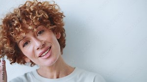 Cute adult curly haired woman twirling hair on finger in the grey studio