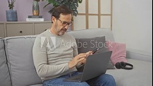 A mature man multitasks with a smartphone and laptop on a couch, indicating productivity in a casual home setting.