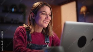Young smiling Caucasian woman in front of laptop in living room. Beautiful blonde using computer to chat online with friends. Millennial female typing on social apps on electronic device at night home
