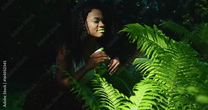 The sensitive outdoor portrait of the afro-american girl with bright green make-up spraying the fern using the green pulverizer in the botanic garden. Green composition.