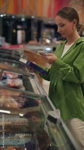 Young woman is comparing products in the frozen food aisle of a grocery store. She is using her smartphone to help her make a decision