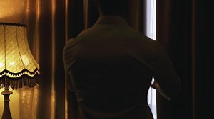 Young man opening curtains at hotel room. Man in white shirt opening the dark window curtains in a room. Relaxed man looking on the window. Back view.