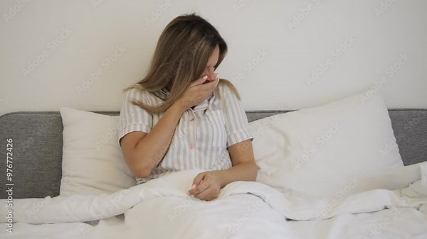 A woman with a cold lies in bed and coughs into a tissue, struggling with a persistent cough. Close-up of the cough and discomfort as she uses the tissue. Severe coughing and illness are depicted.