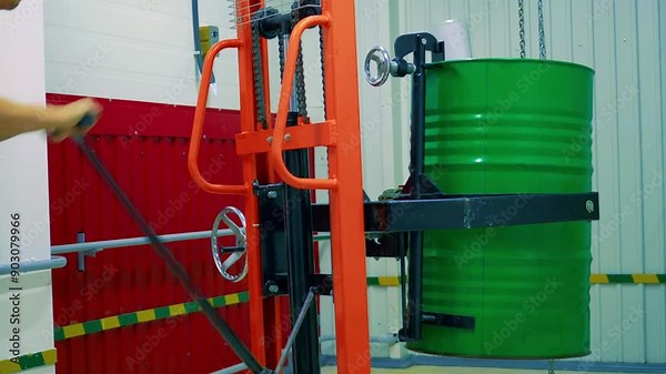 Factory worker in uniform lifts green barrel of chemicals using hydraulic jack. Ensuring safety while transporting hazardous materials. Industrial process showcasing proper handling and transport.