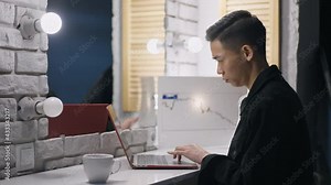 Focused Asian man typing on laptop keyboard analyzing graph in home office at night. Side view of busy concentrated young Chinese expert overworking indoors. Remote working on pandemic lockdown