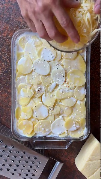 overhead view of person preparing a meal, putting grated cheese onto potato bake in baking tray