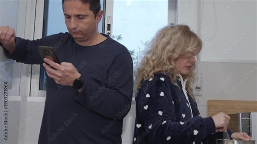 Mature couple having a distant morning routine, man engrossed in his phone while his wife cooks breakfast in the kitchen, feeling neglected and showing indifference