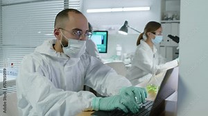 Waist up of Middle Eastern male scientist in protective white coverall and face mask observing liquid in test tube and typing notes into laptop while working in modern lab with colleagues