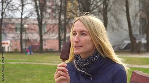 Blonde woman eating ice cream with chocolate icing on a stick. Girl eats ice cream in a park, on a city street.