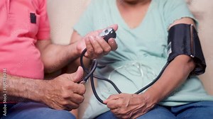 Close up shot of indian senior man checking bp or blood pressure of wife by using sphygmomanometer at home - concept of routine health checkup, medical care and togetherness.