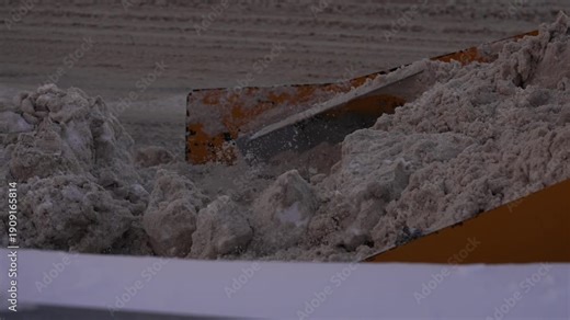 Heavy equipment bulldozer clearing a city road from dirty snow and ice with its front loader bucket, while a municipal service worker with a shovel helps with winter maintenance after a blizzard
