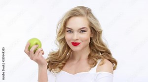 Young beautiful woman eating fruit and says yum. Happy adult girl tastes a green apple. VIdeo footage of a blond caucasian woman eats fruits over white background. 4k resolution.