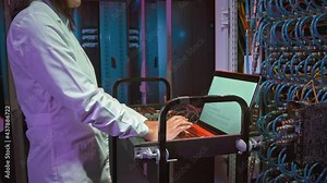 Tilt down shot of female Asian IT specialist in lab coat typing on laptop standing on cart and connected to computing equipment in data center
