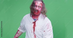 Male disheveled zombie with a bloody face and clothes is staggering and snapping its teeth, wanting to attack and bite its victim, front view, green chromakey on background