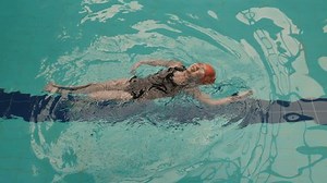 A young girl is learning to swim on her back in an indoor pool. She is floating confidently, wearing an orange swim cap and pink goggles, with her arms spread out for balance.