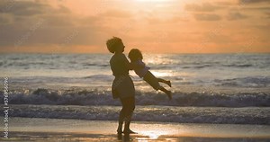 Aunt and nephew play by ocean on beach. The aunt holds and spins boy, having fun and laughing. A woman spins her nephew around while they are on the beach in the warm sunset. Games by the ocean surf.