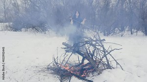 Woman witch near burning bonfire in snowy woodland perform ritual dances in black dress. Ancient mystical spiritual dancing ceremony in winter forest. Scary wizard making ordinance standing on snow.