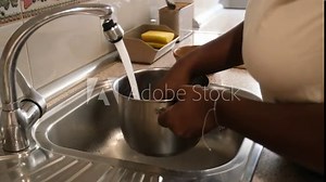 Woman engaged in washing rice in a kitchen sink using a stainless steel saucepan under a running faucet