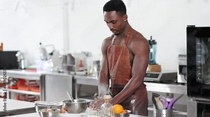 Handsome African American man chef wearing brown leather apron kneading dough on the table. Happy cooking black guy. Making bread, manufacturing process. Workplace, professional kitchen interior.