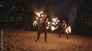 Fireshow artist performing skill of rolling burning dragon staff while two pretty firegirls spinning flaming staves in background. Stylish trio showing art of rolling and spinning lit staves at night