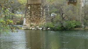 Scene pans down river to a competitive rowing boat emerging rapidly from behind the old stone supports of a bridge.