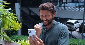 Handsome dark-haired bearded Caucasian young man using smartphone, scrolling news feed and checking social media content, clenching fists from happiness, expressing surprised emotion from good news