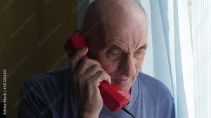 cheerful caucasian pensioner talking on the phone while standing at the window with blinds. lifestyle of the elderly. wrinkled face of an old man with a telephone receiver