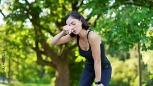 Young brunette female runner is short of breath after a fast sprint run in urban city park. Fit female athlete leans on her knees, wipes the sweat from her forehead with her hand and breathes deeply