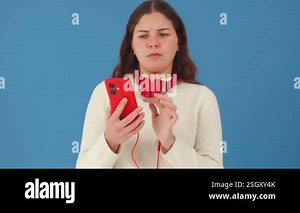 Young woman with red headphones around her neck holding a smartphone and making a confused expression. She appears puzzled or frustrated, looking away. Solid blue background Stock Video Footage - Alamy
