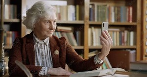Caucasian gray-haired old beautiful woman in glasses posing to camera of smartphone while sitting at table in library. Female librarian taking selfie photos with phone at desk in bibliotheca.