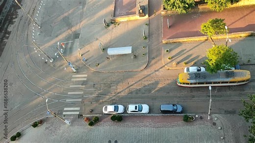 Passing tram at road intersection. Road, tram, people, parked cars, green trees and shadows on summer morning. Shining sun. Cityscape. City scenery. Town landscape. Top view. View from Above