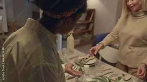 Tilt up over the shoulder view of African American woman serving table with Caucasian mother-in-law while preparing to have dinner with family