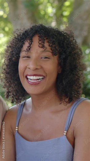 Vertical video: Laughing woman turning under tree while friend teasing, wearing dress and clip mic