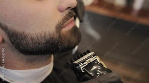 Man shaving customer beard with a trimmer. A master hairdresser with a professional trimmer in a black gloves makes a haircut and evens the beard to a customer.
