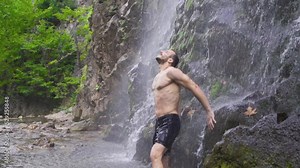 Strong male standing under waterfall. Slow motion. Sporty strong man getting wet under waterfall.