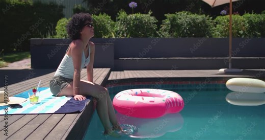 Woman is lowering feet into pool after touching water, nudging pink float and relaxing on deck