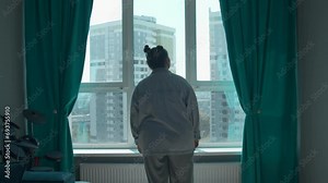 Young woman enjoying view through the panoramic window in the morning at home. Media. Girl in pajamas standing and thinking by the window in bedroom.